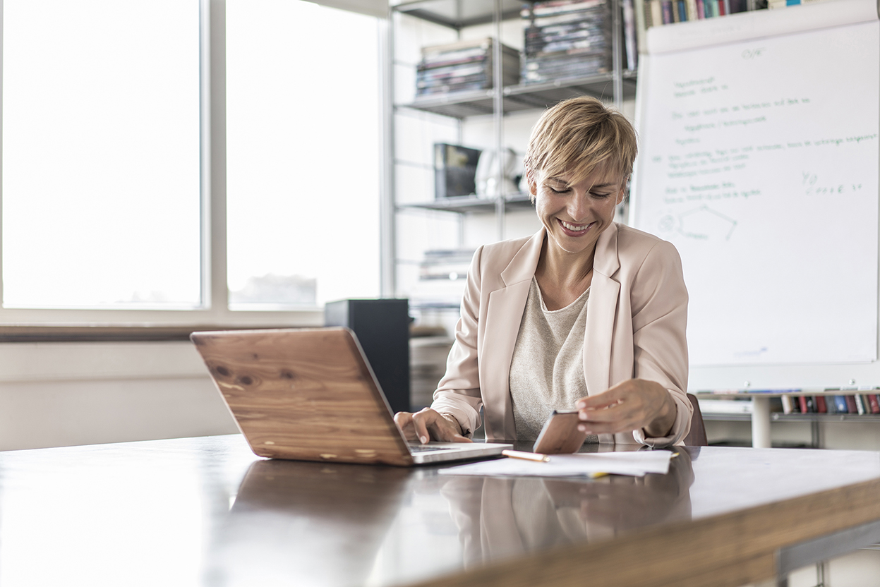 Smiling businesswoman with cell phone and laptop in modern conference room